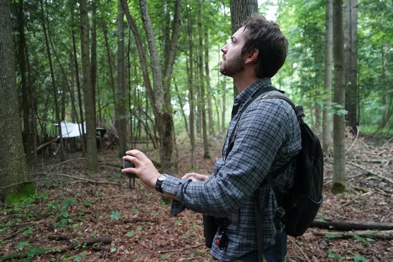 A man in a forest hold a small Bluetooth speaker in his hand.