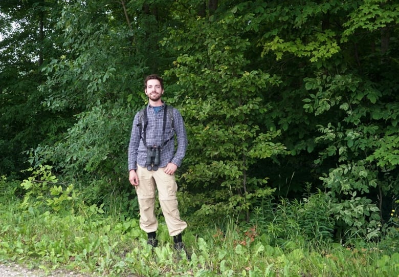 A man with binoculars stands in front of a forest. 