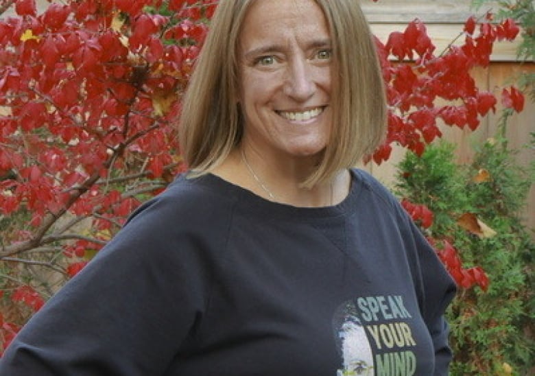 A woman with short blond hair, wearing a black sweatshirt, smiles while she poses for a portrait in front of a red-leaved tree.