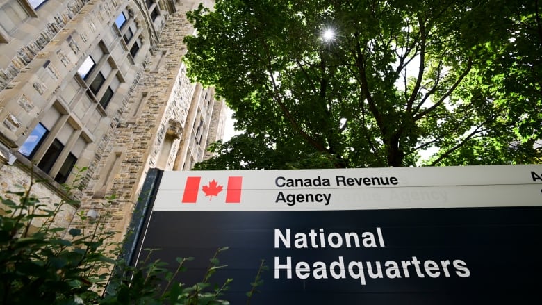 A sign reading "Canada Revenu Agency" under the shade of a tree stands next to an old brick building.