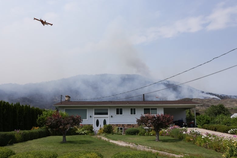 A water bomber flies over a white rancher as a wildfire burns on a brown, desert hill in the distance. 