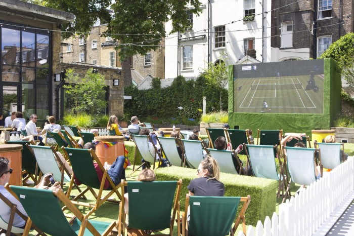 Spectators in deck chairs watching the tennis on a large screen at Bluebird Chelsea