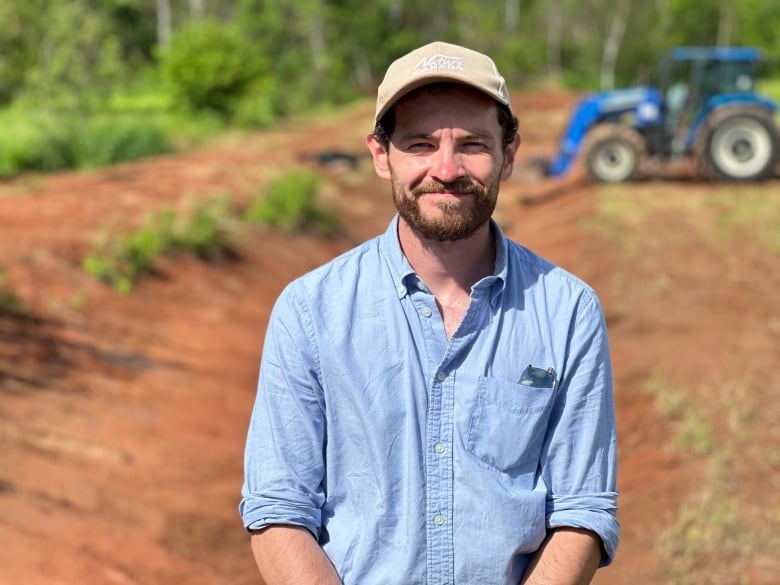 A man in a blue shirt and ballcap stands near a tractor and ditch 