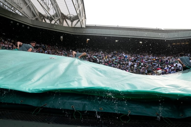 Two ground staff pull a large tarp over the court as rain falls.