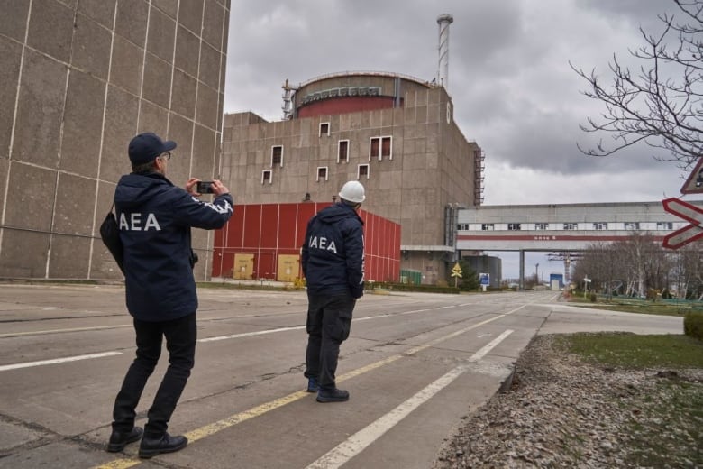 A man wearing a blue jacket with the letters IAEA, for International Atomic Energy Agency, on the back, takes a photo of another person in a similar jacket and white hardhat at a nuclear power plant in Ukraine.