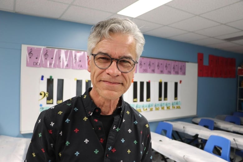 A man smiles at the camera. Behind him is a classroom with a whiteboard. 