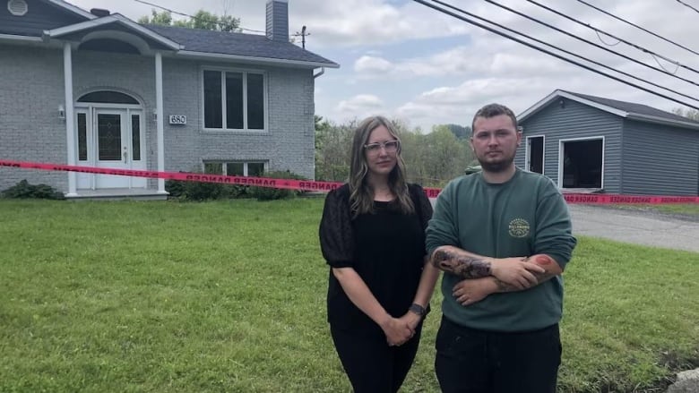 A young woman and man stand in front of a house with grey siding, with a nearby matching shed. There is red caution tape between them and the house.