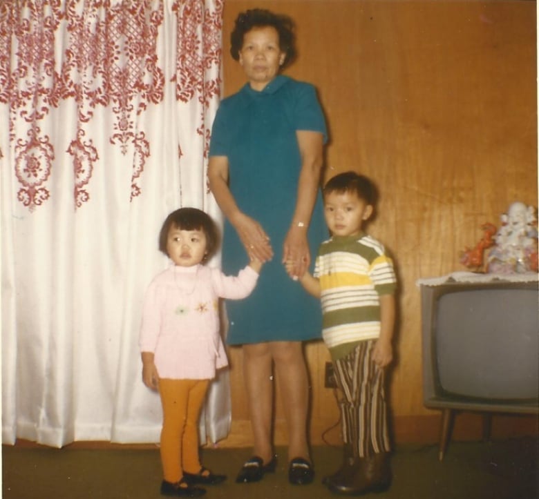 A woman with a perm stands in a room beside an older model television. She's holding the hands of two children. 