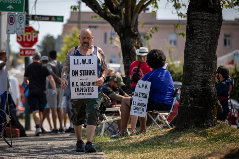 A man stands with an "On Strike" sign hanging from his neck on a sidewalk, with a group of picketers in the background. 