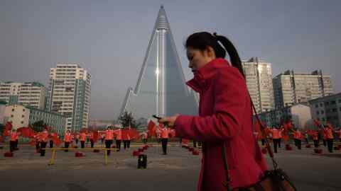 A woman on her phone passes a propaganda dance troupe in front of the Ryugyong hotel in Pyongyang, North Korea