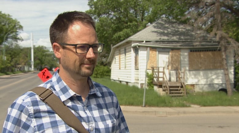 A white man with short dark hair, a short beard and glasses is wearing a blue plaid button-up short sleeve shirt. He is standing on a street corner. Behind him to the left are two orange construction signs; behind him to the right is a white house with grey shingles, plywood over the windows and unkempt grass.