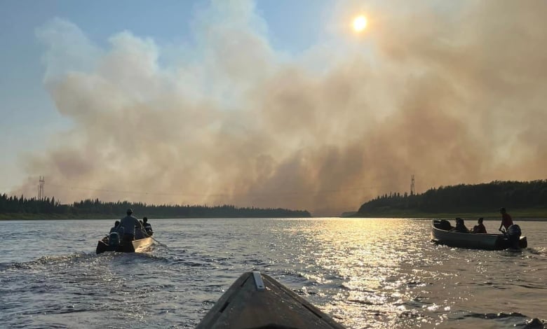 Boats make their way across a wide river with large plumes of smoke obscuring the sun