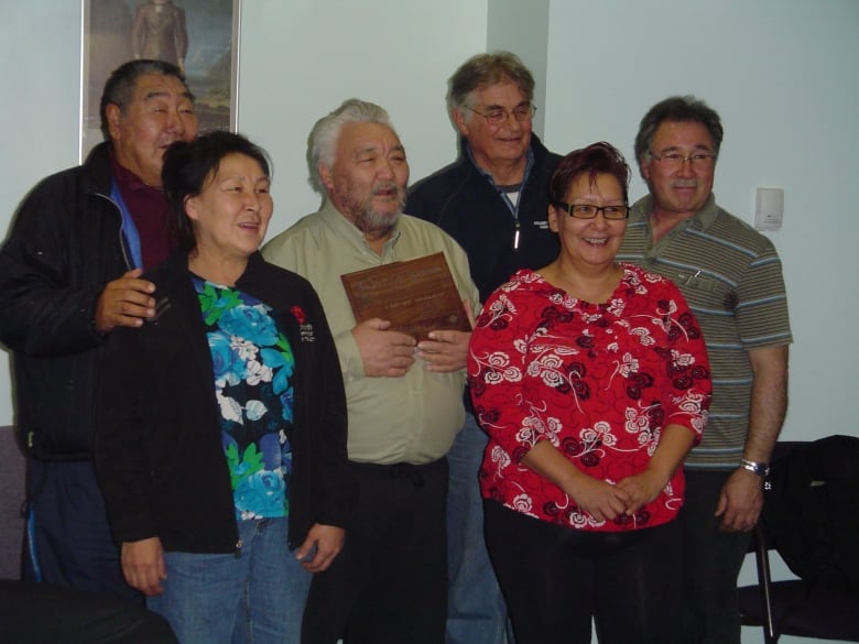 Six people stand in a group posing for a photograph, with the man in the centre of the group holding a plaque.