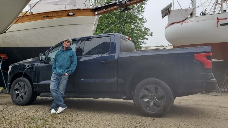 A wide shot showing Ian with his back to the driver's door of his truck. The charger is plugged into the vehicle to his right.  The hull of a large sailboat and another boat are seen in the background.