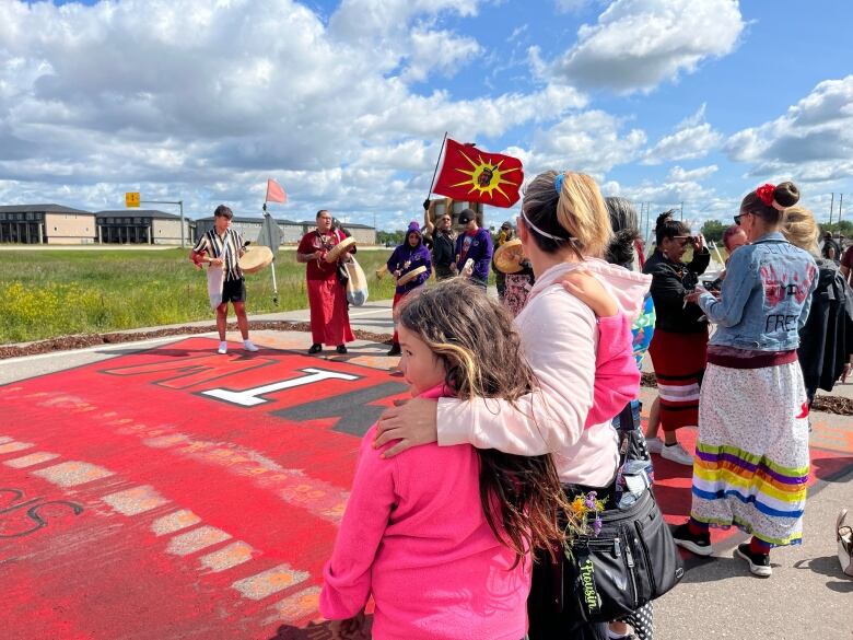 A group of people stand around a painting on the ground of red dress that says "MMIW." Some of the people hold red and yellow flags, while others are holding hand drums.