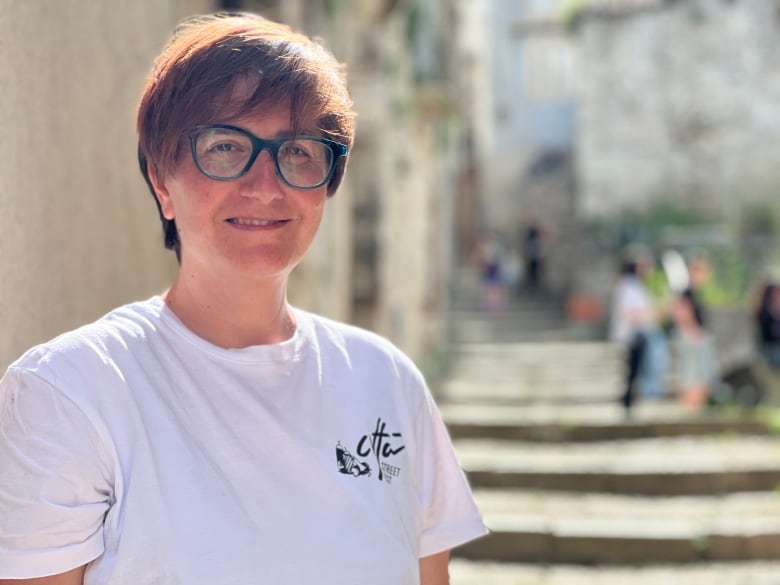 A woman with short brown hair and black rimmed glasses, wearing a white T-shirt, poses for a portrait in front of a set of stone steps.