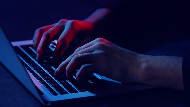 A closeup shows a man's hands typing on the keyboard of a laptop computer in a darkened room.