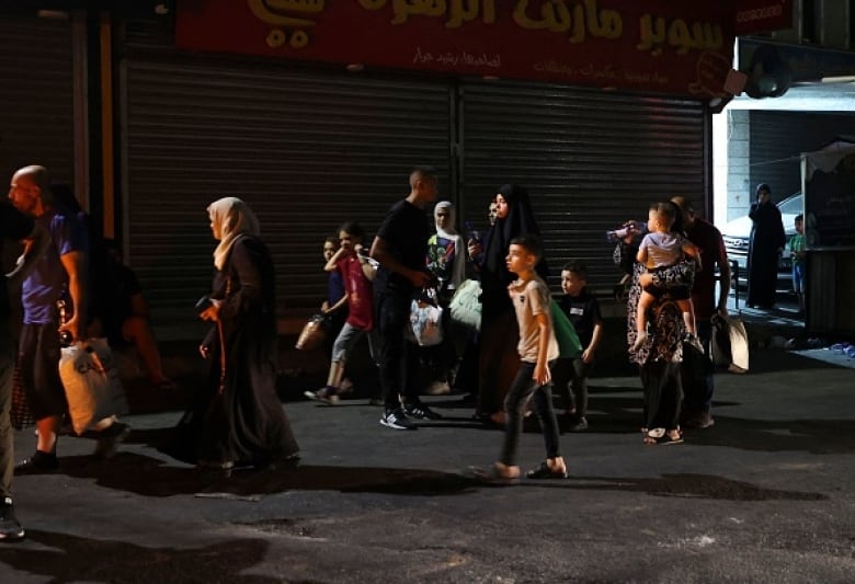 Men, women and children walk on a street at night. Some carry bags that appear to contain clothes. One woman carries a small child.