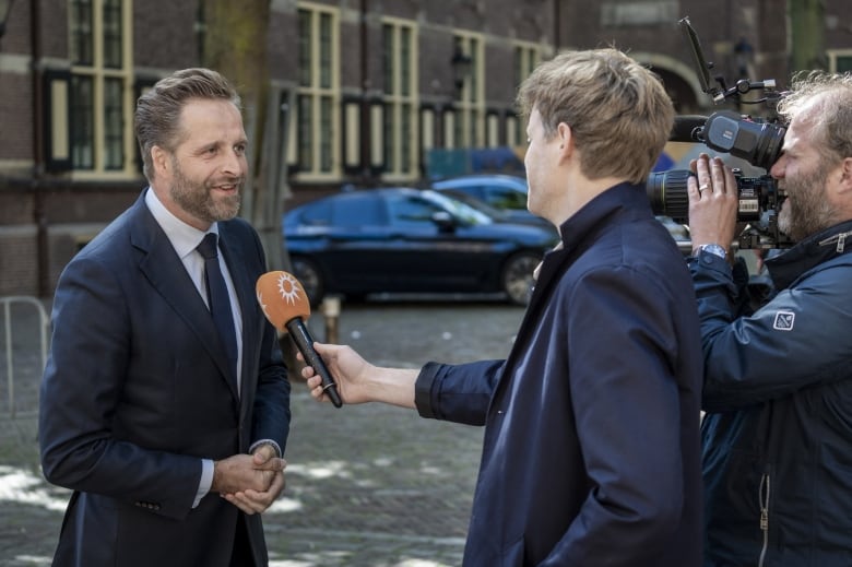 A man in a suit speaks to a reporter holding a microphone while a cameraman records them. 