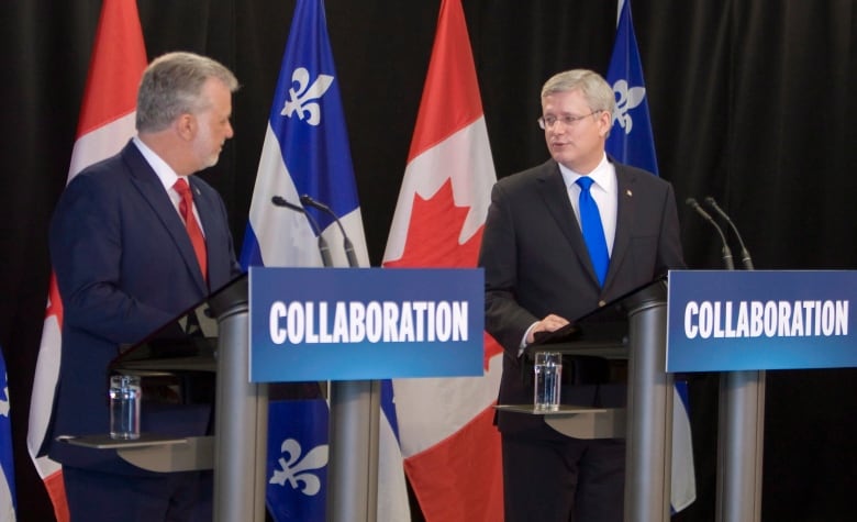 Two men in suits stand at podiums in front of flags.