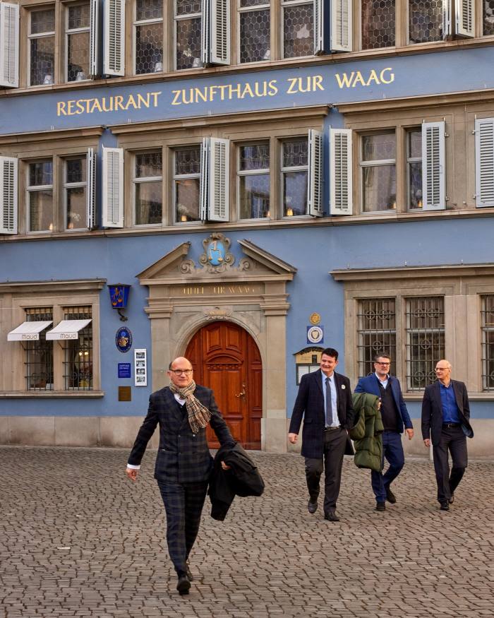 Men in suits walking across cobbled paving in front of the blue and grey facade of the Zunfthaus zur Waag 