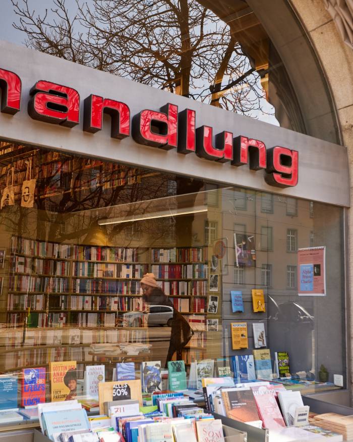 Rows of books on table in the front of Buchhandlung im Volkhaus; through the window is a female customer browsing shelves of books