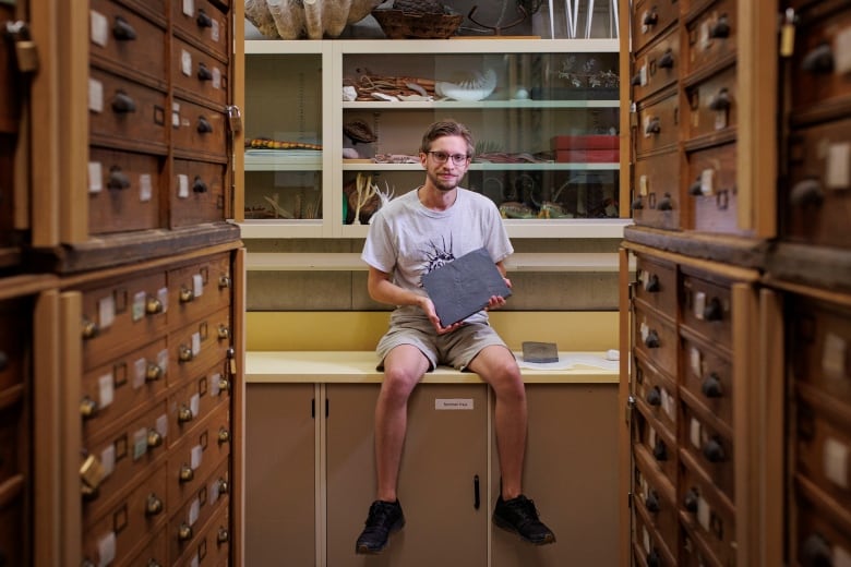 A man sits on a lab bench holding a slab of dark stone at the end of an aisle of wooden drawers on either side.