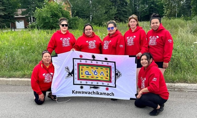 A group of seven women in red sweatshirts hold the flag for Kawawachikamach. 