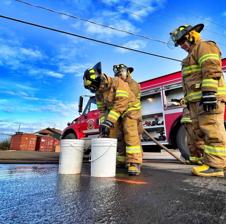 Women in firefighting gear bend over a bucket near a firetruck 