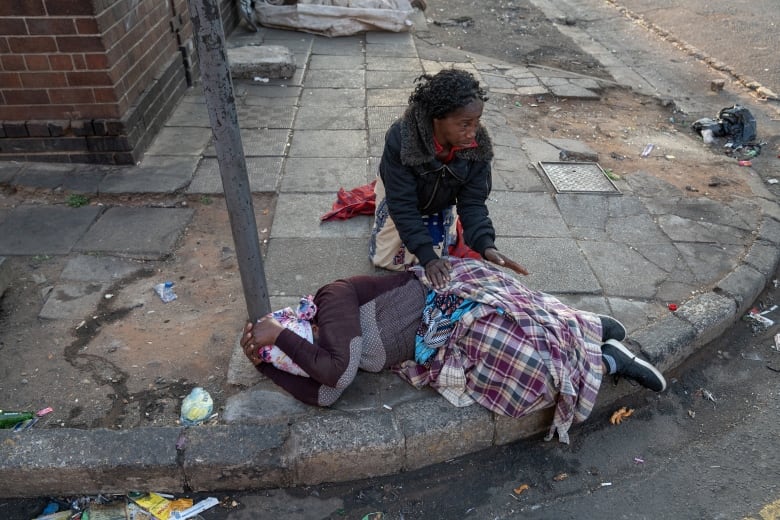 A woman lies on her side on a sidewalk holding her head in her hands. Another woman kneels behind her, with both hands on her back.