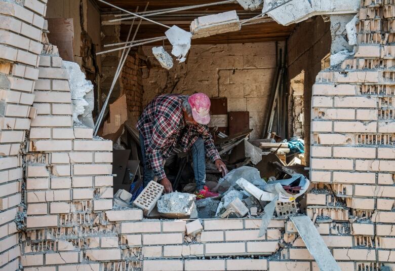 A person in a red flannel top and dusty red baseball cap searches through the ruins of a brick building damaged in an explosion.