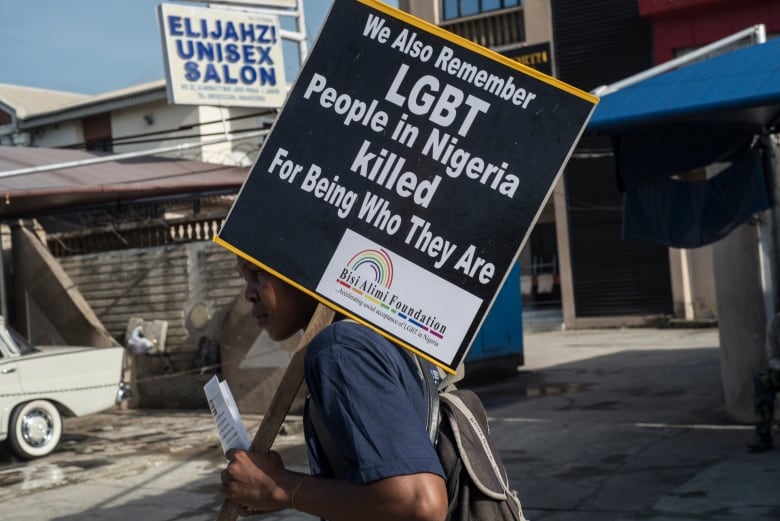 A man holds a protest sign over his shoulder that reads "We also Remember LGBT People in Nigeria killed For Being Who They Are." 