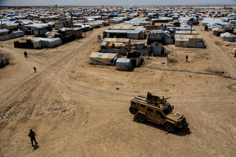 Make shift shelters dot the horizon as a military truck drives by in the foreground.