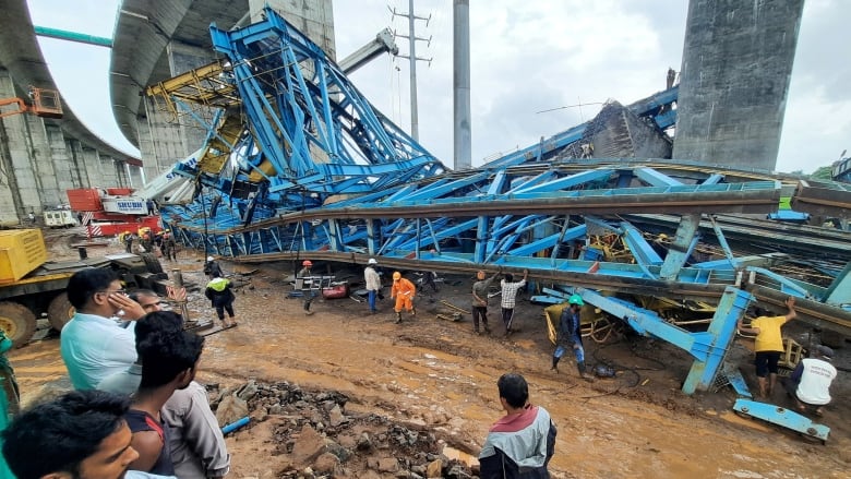 A massive blue crane is shown downed on the dirt, with several people shown walking near the site, located near an overpass.