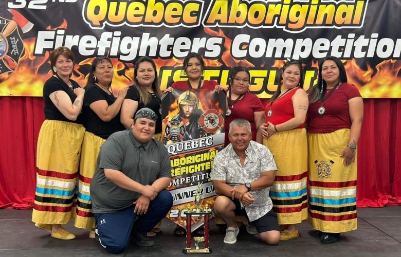 A group of women stand on a stage with two men with a trophy. Behind them hangs a banner that says Quebec Aboriginal Firefighters Competition. 