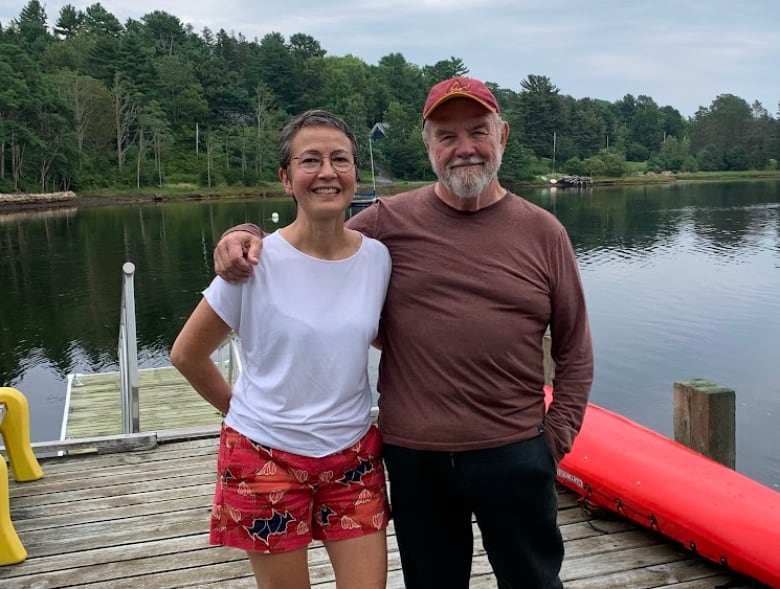 A couple, arms around each other, stand on a dock on a lake.