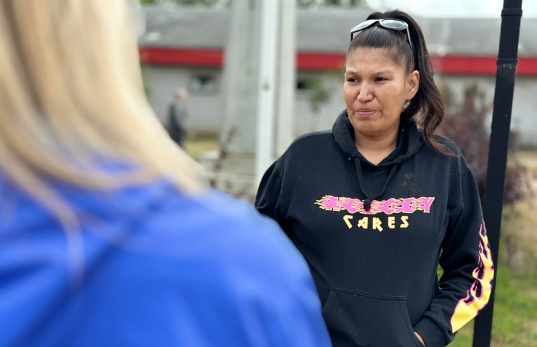 A woman wearing a hoodie and sunglasses on top of her head looks off to the left. In the foreground is a woman wearing a blue jacket.