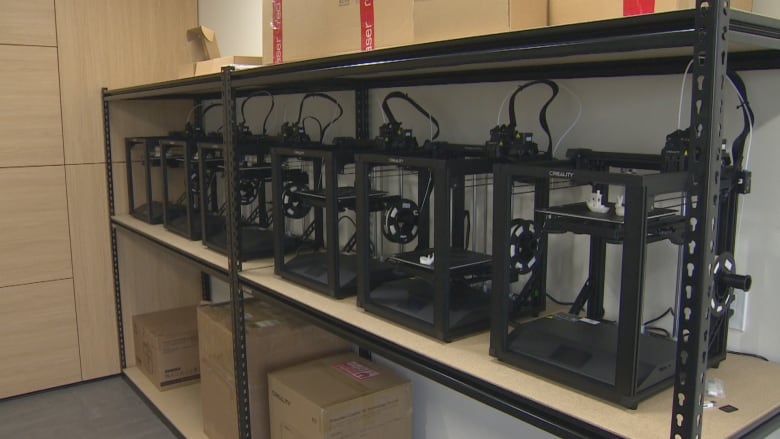 Black 3D printers are on a shelf in a room.