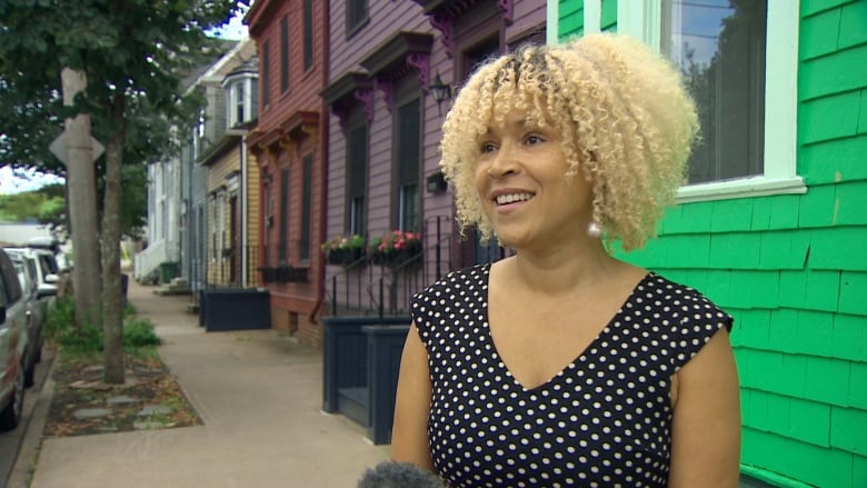 A Black woman with curl blonde hair wears a black and white polka dot dress, standing in front of a bright green older-looking home