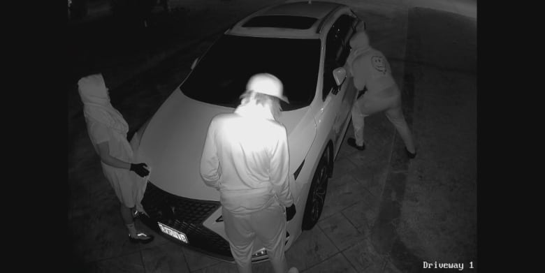 A screenshot from video shows three people wearing hooded clothing and gloves attempting to steal a car from a driveway. 