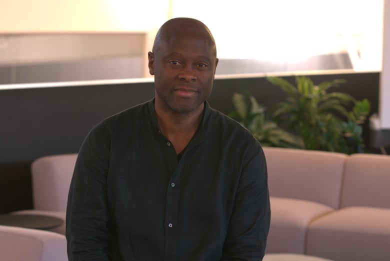 A Black man in a black shirt, unbuttoned at the collar, smiles at the camera while seated in front of a tan sofa. 