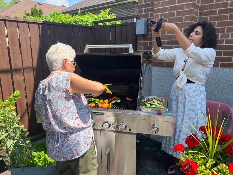 A woman with white hair, wearing a flowered blouse and apron, stands on a patio with her back to the camera, grilling vegetables on a barbecue as a young woman with curly dark hair watches and holds a camera and microphone up to record. 