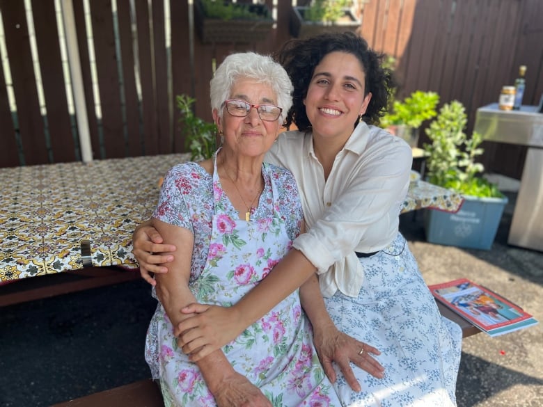 An older woman with short, white hair and glasses and wearing a flowered shirt and apron, is hugged by a younger woman with long, curly black hair. 