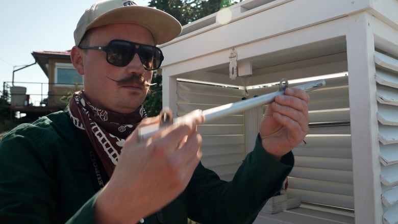 A man wearing a cap, dark sunglasses, a dark shirt and dark red scarf is seen examining a tool.