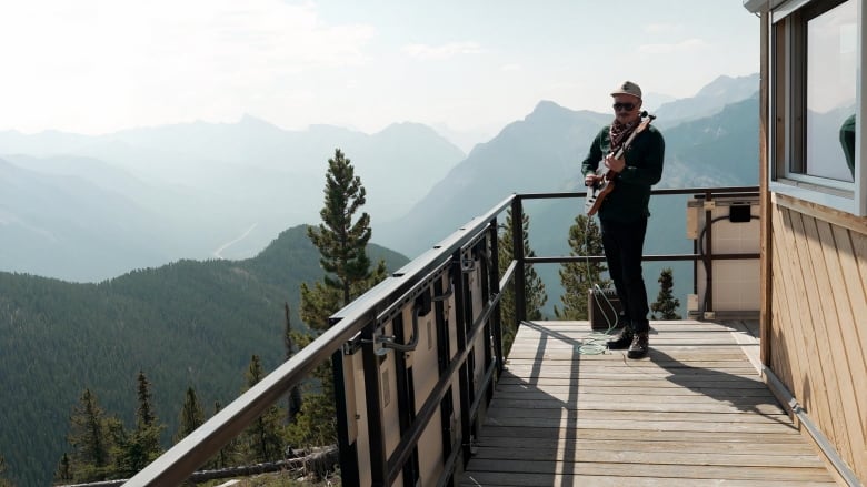 A man is seen playing a guitar on a balcony. Mountains and trees are visible in the background.