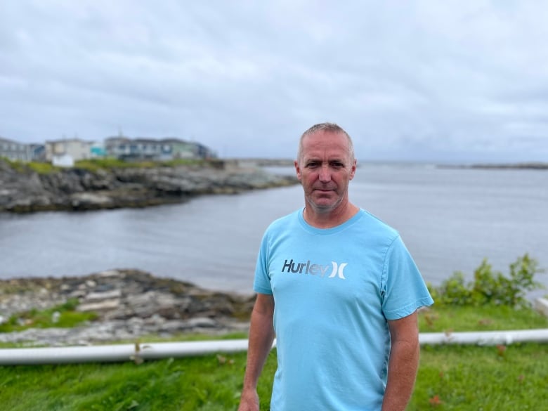 A man in a light blue t-shirt with the word "Hurley" on it stands against the shore in Channel-Port aux Basques, N.L.