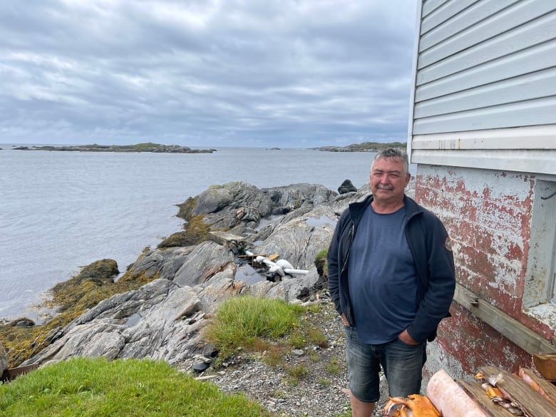 A man in a blue shirt, a darker blue windbreaker and jean shorts stands across from the coastline in Channel-Port aux Basques, N.L.