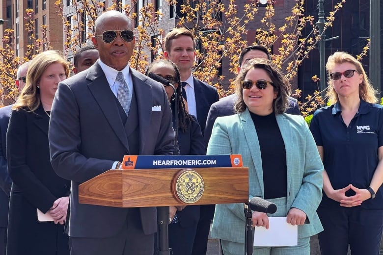 A bald man in a suit and sunglasses stands at a podium outside next to a woman, also in a suit and sunglasses. Several people stand behind them. 