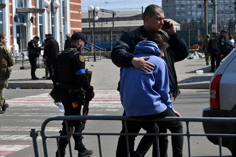 Man speaking on a phone with his arm around a person wearing a blue hoodie, sitting on a railing