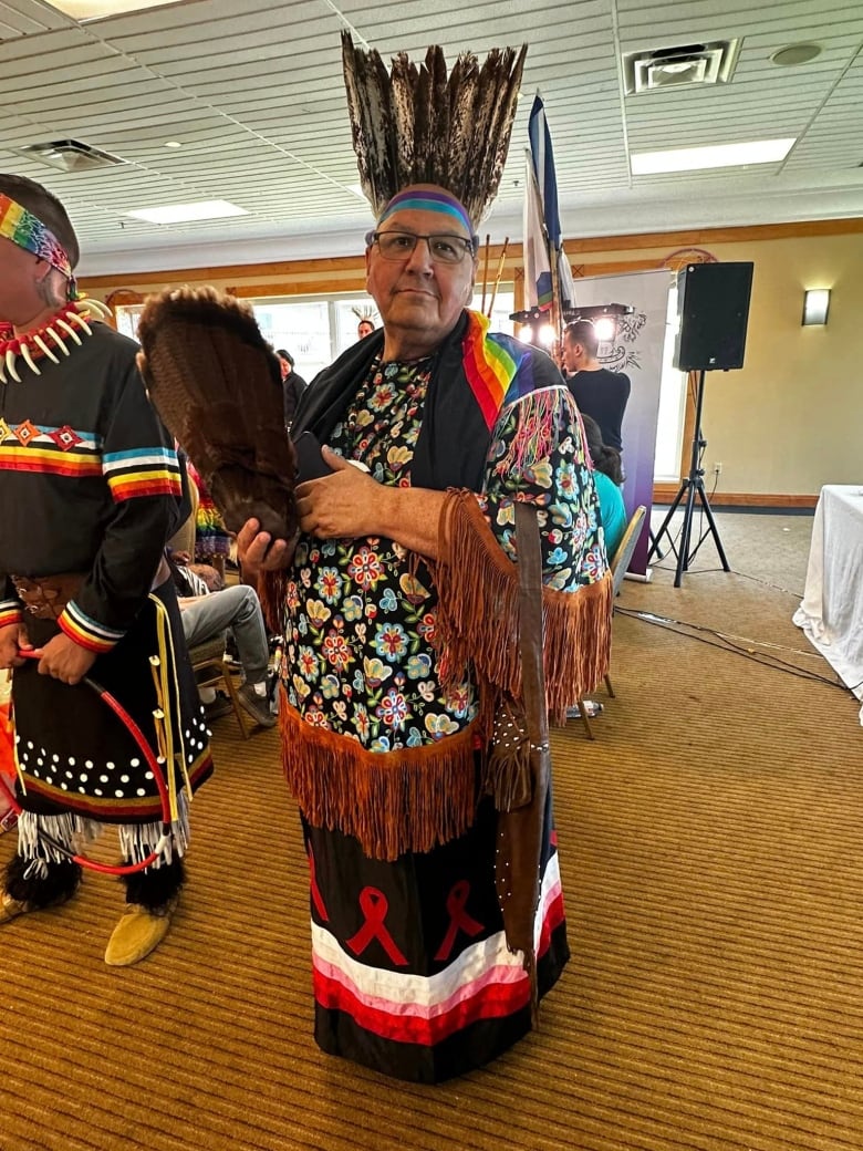 An older person is dressed in a black ribbon skirt decorated in white, pink and red, with a flower-patterned top trimmed with brown fringe. A rainbow sash is draped on their shoulders. They're wearing a feather headdress and carrying an eagle wing.